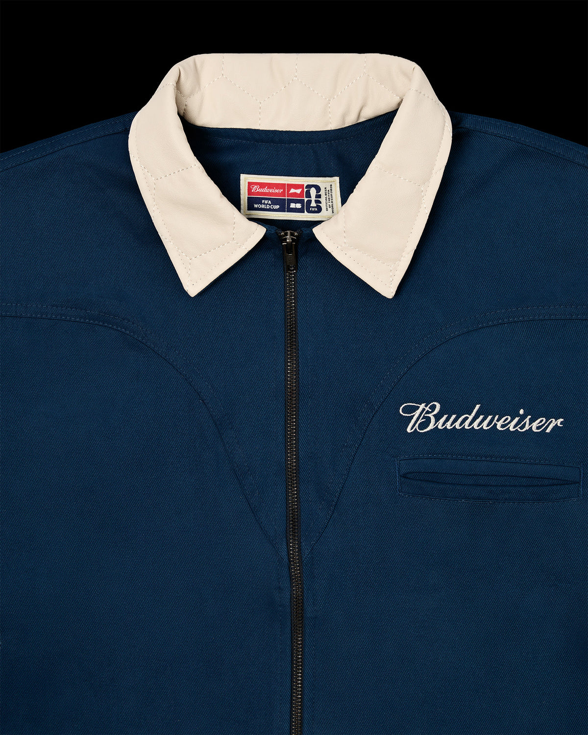 Detalhe da Jaqueta Worker Kiji Studio para Budweiser Copa do Mundo 2026, na cor azul, logo 'Budweiser' bordado em branco e gola em couro texturizado.